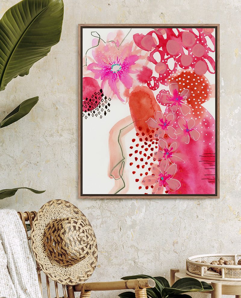 A framed abstract floral art print with pink and orange colors hanging on a wall, with a straw hat and plant in the foreground.