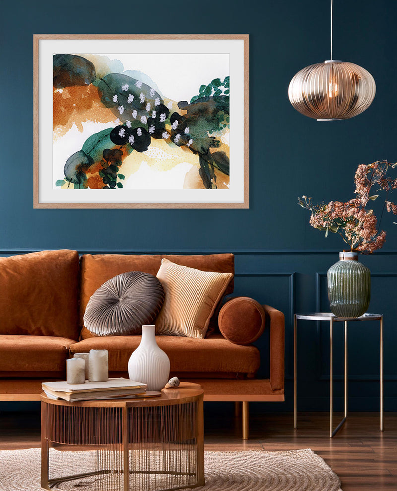 A framed abstract artwork with flowing green, brown, and orange elements displayed on a wall above a brown sofa with decorative pillows, a white vase, and a wooden coffee table in a room with a dark blue wall and a hanging light.