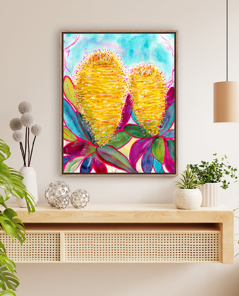 A vibrant abstract floral art print featuring shades of yellow, magenta, and blue displayed on a table with decorative items and a wall lamp in the background.