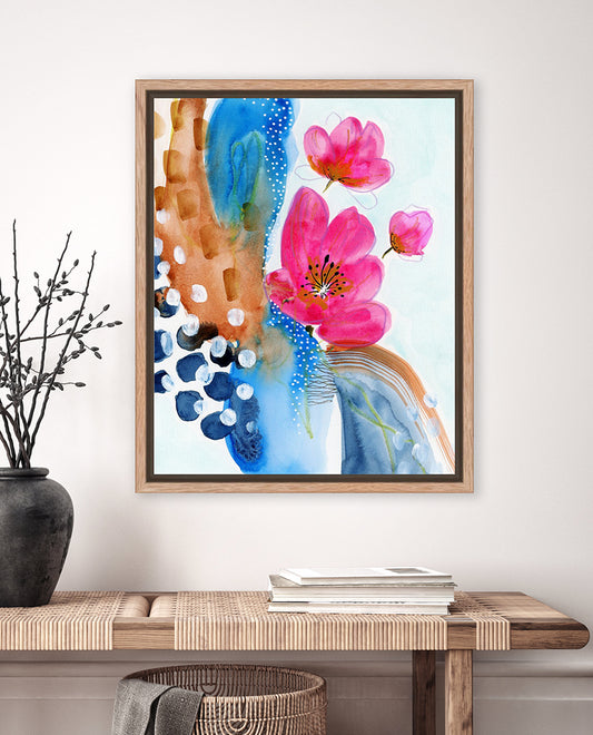 A framed abstract floral artwork featuring pink petals and deep browns and rich blues on a wall, with a wooden surface below.