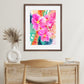 A framed art print featuring vibrant pink floral artwork with corals, gold, emerald green, sapphire blue, and orange accents, displayed on a wall above a wooden table and chair.