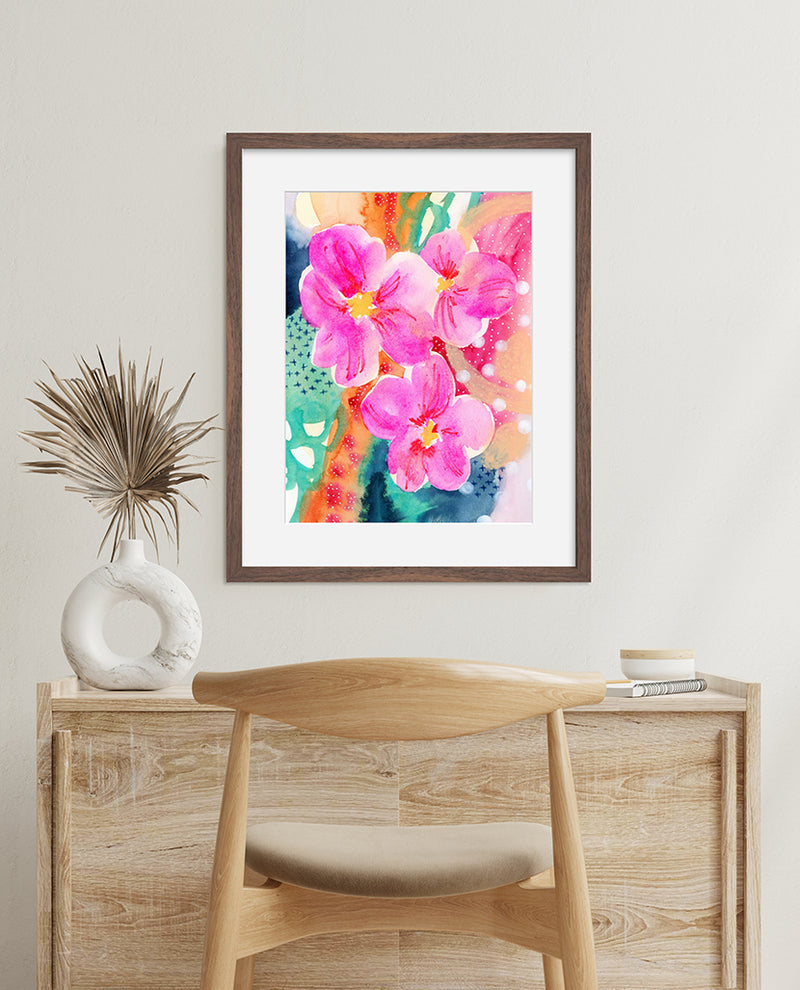 A framed art print featuring vibrant pink floral artwork with corals, gold, emerald green, sapphire blue, and orange accents, displayed on a wall above a wooden table and chair.