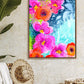 A vibrant floral artwork titled 'Joyful Garden' displayed on a wall above a tropical hat collection, with pink and blue flowers and leaves on a white background.