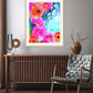 A vibrant abstract floral art print with fiery pinks, radiant oranges, and deep crimsons, titled 'Joyful Garden', displayed in a room setting with a wooden console table and a modern armchair.