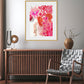 A framed abstract floral artwork titled 'Juney' displayed on a wall above a wooden console table, with a red and pink color scheme, and a round flower vase with a branch on the table.