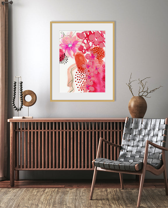 A framed abstract floral artwork titled 'Juney' displayed on a wall above a wooden console table, with a red and pink color scheme, and a round flower vase with a branch on the table.