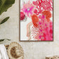 A framed abstract floral art print with pink and orange colors hanging on a wall, with a straw hat and plant in the foreground.