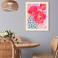 A framed abstract florals artwork with bold pink and red flowers and deep blue accents, displayed on a wall in a room with wooden furniture and a hanging rattan light fixture.