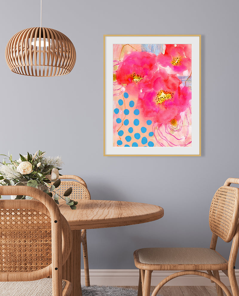 A framed abstract florals artwork with bold pink and red flowers and deep blue accents, displayed on a wall in a room with wooden furniture and a hanging rattan light fixture.