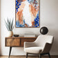 A framed abstract artwork featuring earthy and cool tones with circular patterns, displayed on a wall above a wooden side table and a beige armchair.