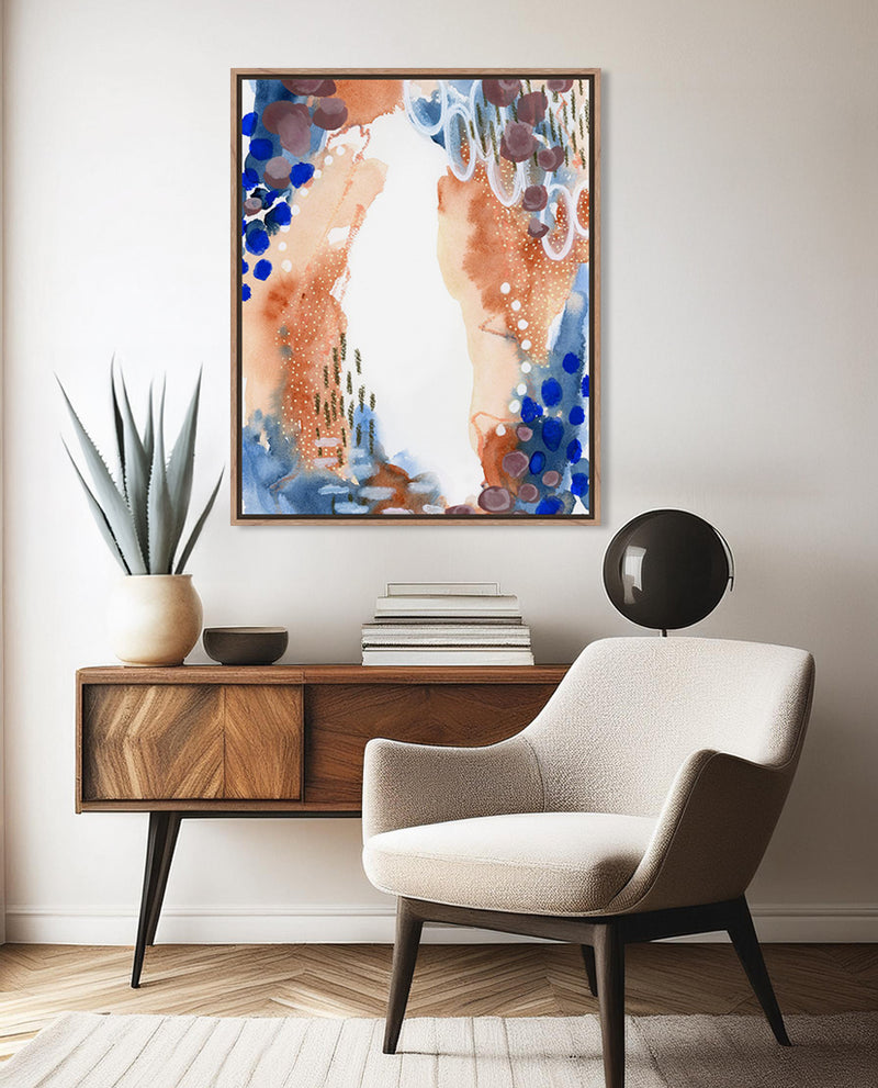 A framed abstract artwork featuring earthy and cool tones with circular patterns, displayed on a wall above a wooden side table and a beige armchair.