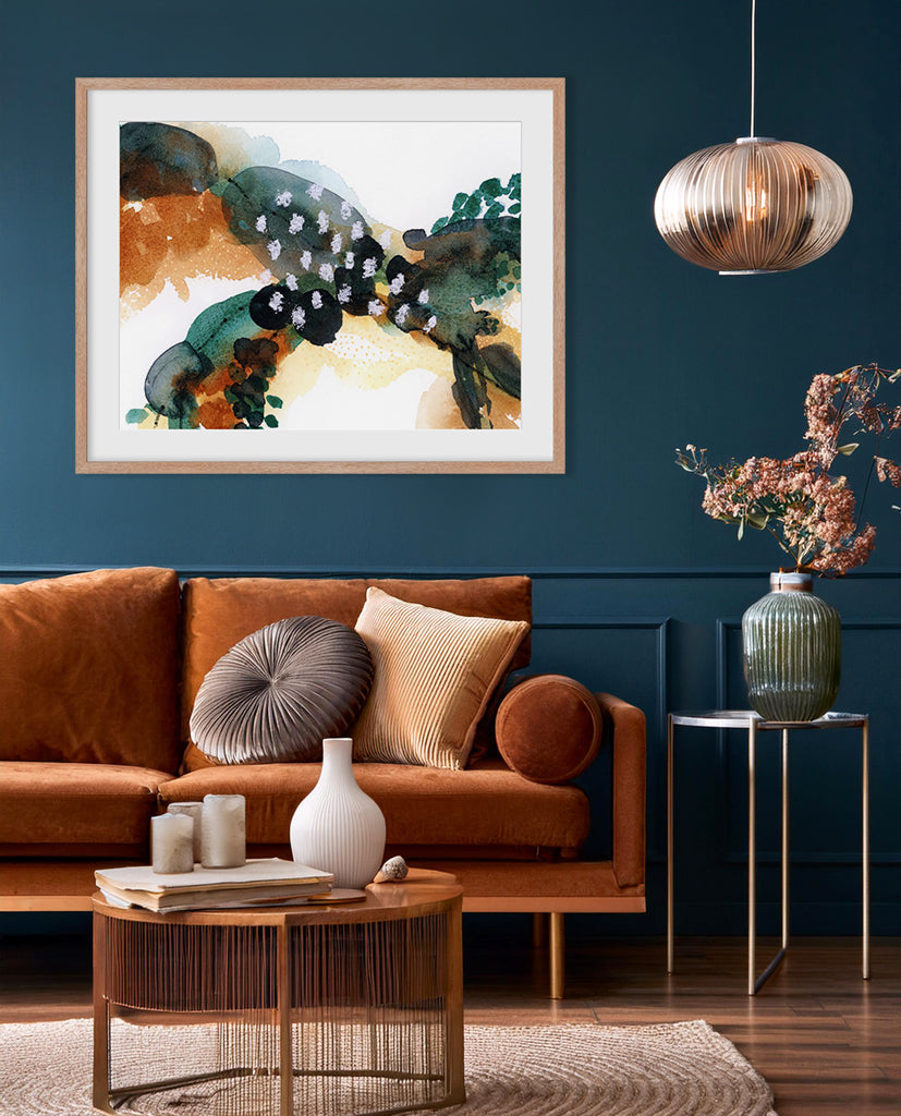 A framed abstract artwork with flowing green, brown, and orange elements displayed on a wall above a brown sofa with decorative pillows, a white vase, and a wooden coffee table in a room with a dark blue wall and a hanging light.